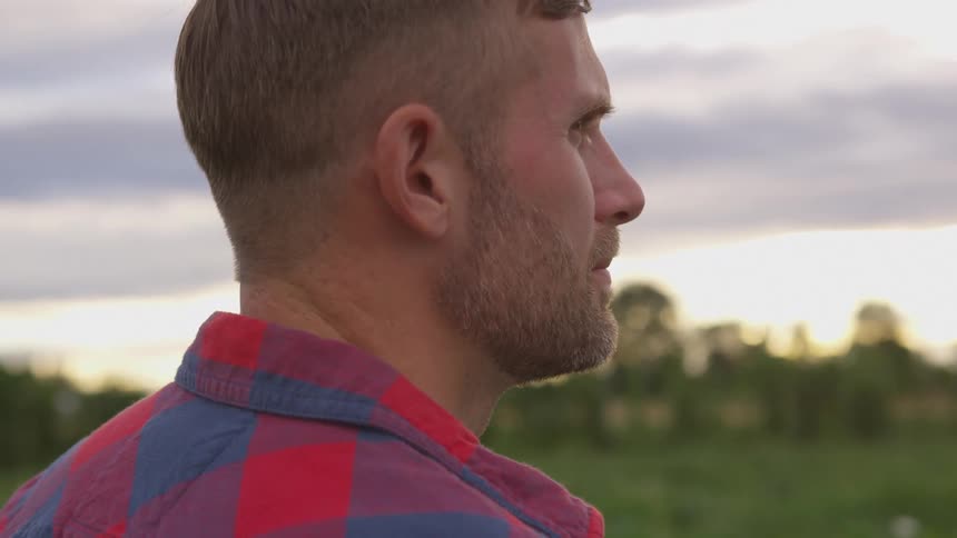 A young man with short hair and a beard wearing a red and blue plaid shirt looks to the right against a blurred background of a field and a cloudy sky