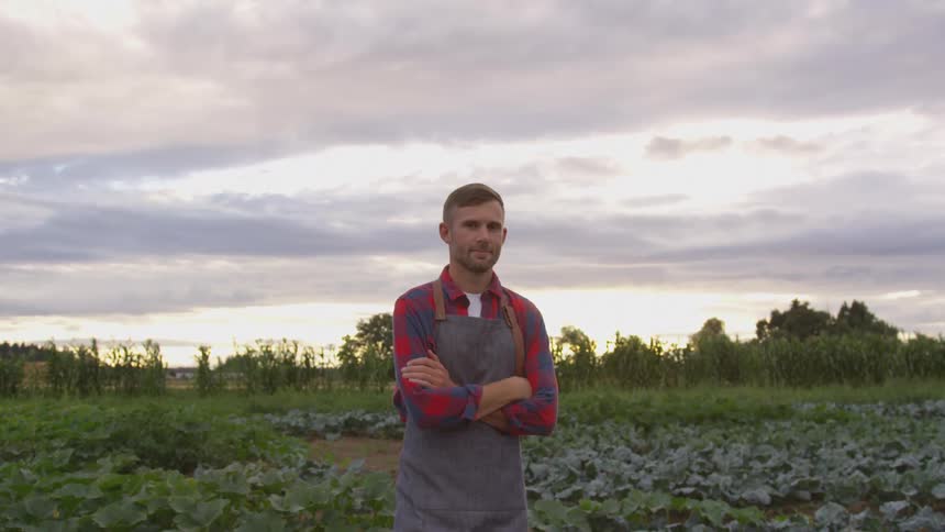 A young man with a beard and short hair stands with his arms crossed in a field of crops wearing a red plaid shirt over a white tshirt and a grey apron under an overcast sky