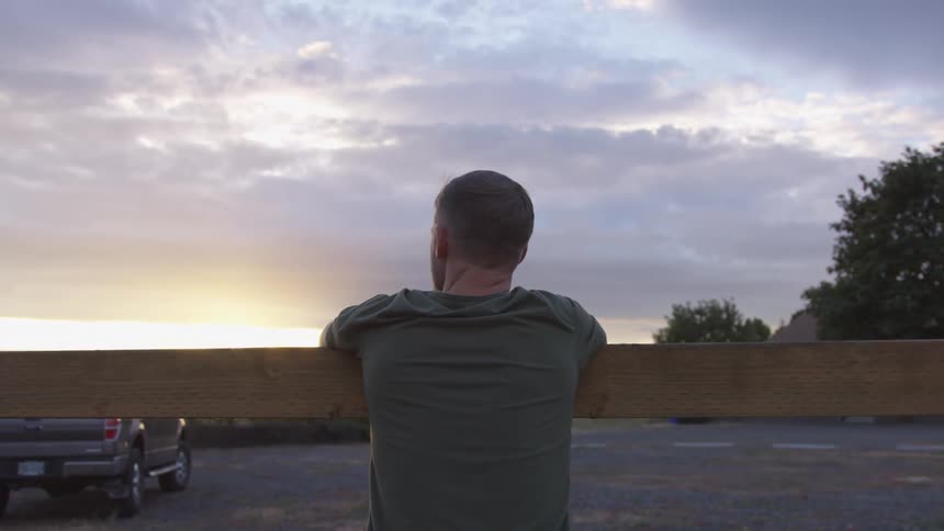 Man stands outdoors at dusk staring into the sky while leaning on a horizontal plank