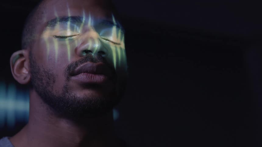 A man with his eyes closed and a beard has his face illuminated by vertical stripes of light in a dark setting