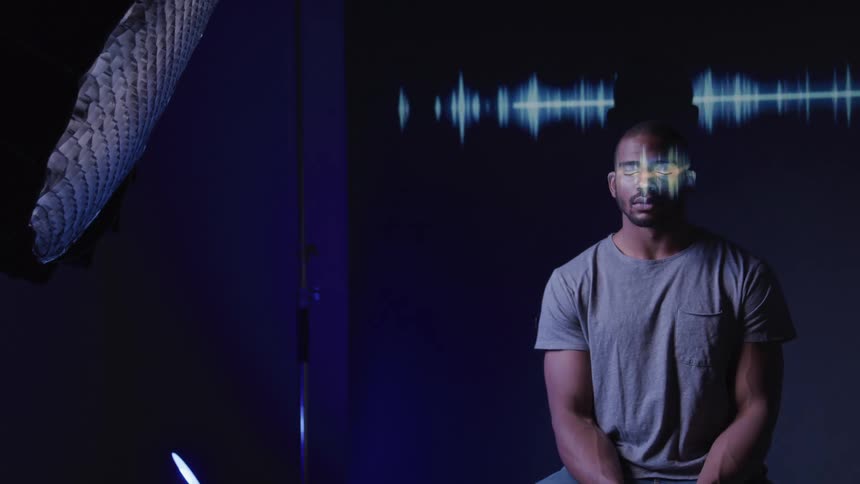 A man in a gray tshirt sits with a reflective light pattern from a bright blue sound wave graphic projected onto his face and the dark background