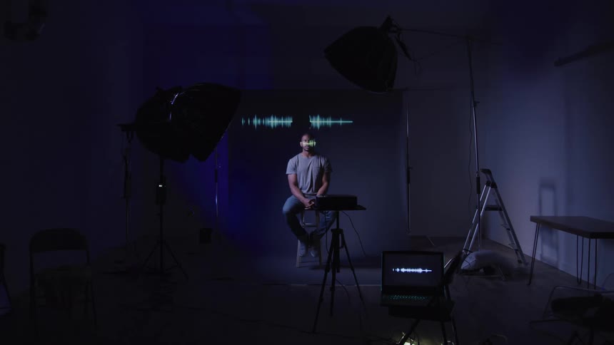 Young black man sits in a photography studio under studio lights with wavelengths projected from a laptop displaying above him
