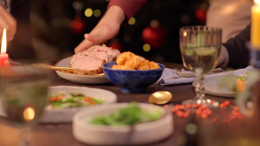 A dinner table set with various meals and a glass of wine as one person places two plates of food on the table while another picks up a spoon beside a plate ready to eat