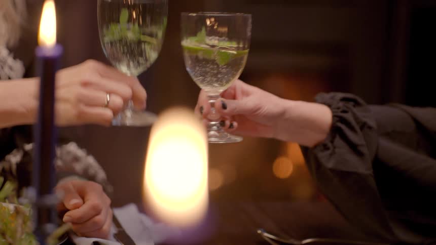 Two hands hold and clink clear glasses filled with a liquid and garnished with lime and mint illuminated by candlelight