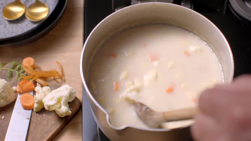 A chef stirs a pot of soup on a stove in a kitchen using a wooden spoon adds an ingredient then lifts the pot off the stove