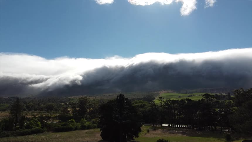 A thick white cloud formation hangs low over a distant mountain range with green trees and scattered buildings in the foreground under a blue sky