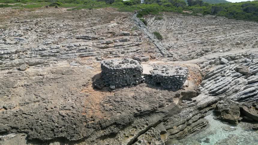 Two rough stone walled buildings sit on a rocky coast on a summer day
