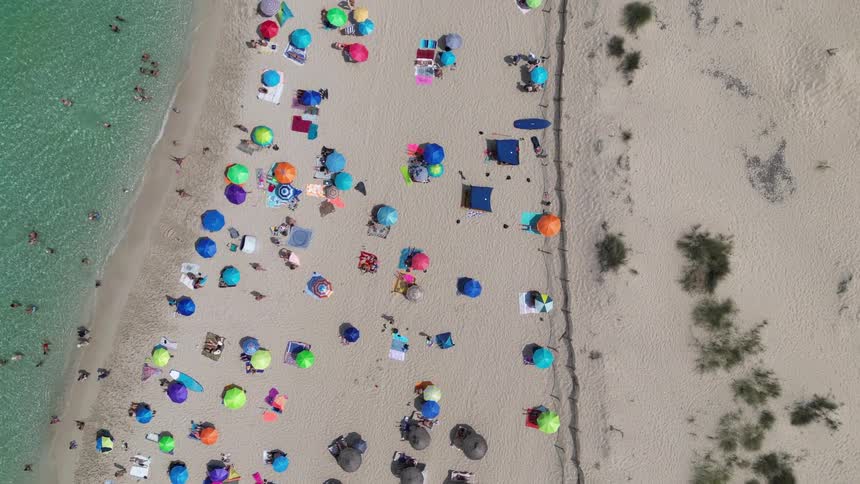 An aerial shot captures a crowded sandy beach with numerous colorful umbrellas and sunbathers next to turquoise water with people swimming in the ocean