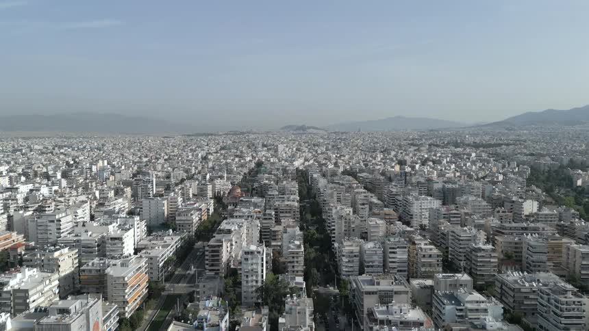 A city skyline featuring numerous skyscrapers and highrise buildings at midday with a coastal road below as a series of cars drive by