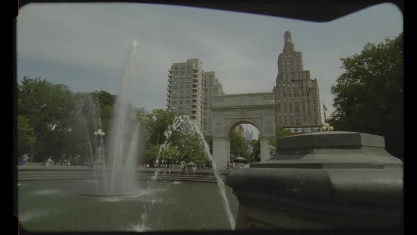 A fountain sprays water into a pool in a park with a large stone arch tall buildings and trees under a cloudy sky