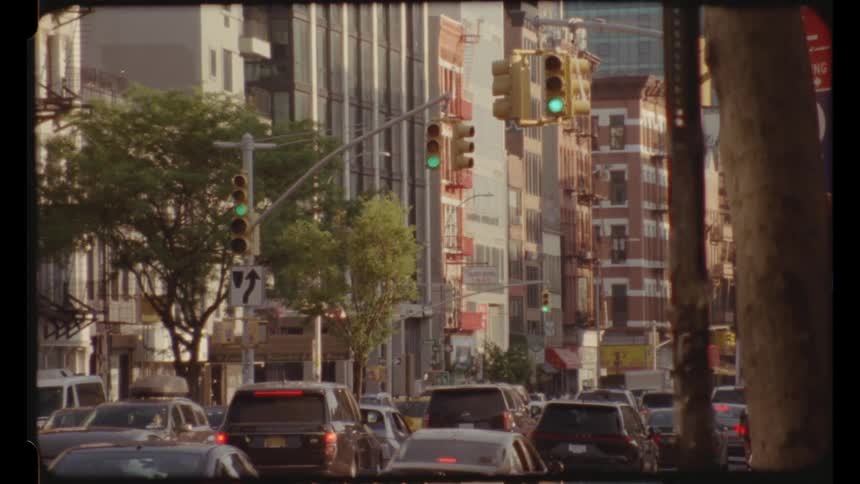A view framed by a tree trunk shows a busy city street with multiple cars a yellow taxi and a bus with buildings and traffic lights under a bright sky