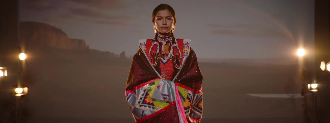 A teenage girl from a native american tribe wears full traditional attire while standing on a stage