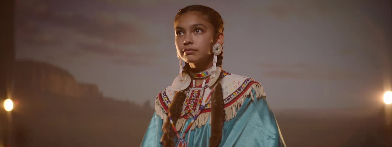 A young girl with braided hair wears traditional native american regalia standing in front of a projected desert landscape with studio lights on either side