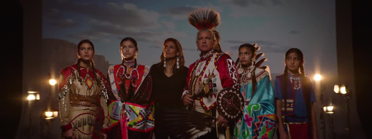 A group of people dressed in traditional native american attire stands in front of a scenic backdrop with professional lighting equipment surrounding them