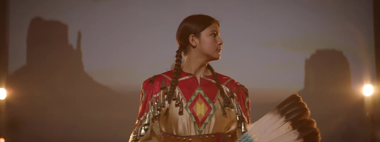 An american indian woman dressed in traditional attire stands confidently on a stage proudly displaying her cultural outfit