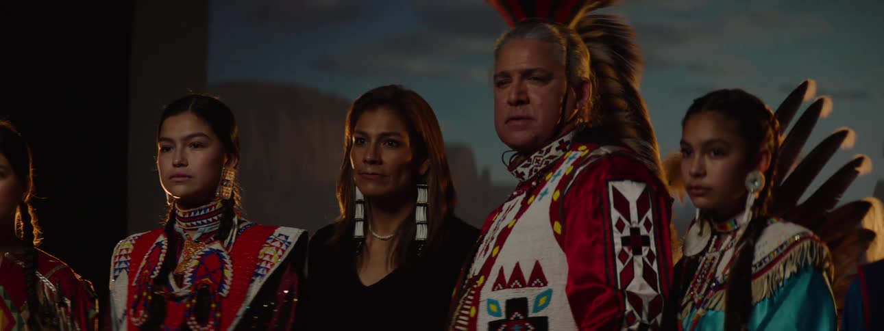 A group of indigenous people some in traditional regalia and others in contemporary clothing with traditional accessories stand and look intently towards the left of the frame