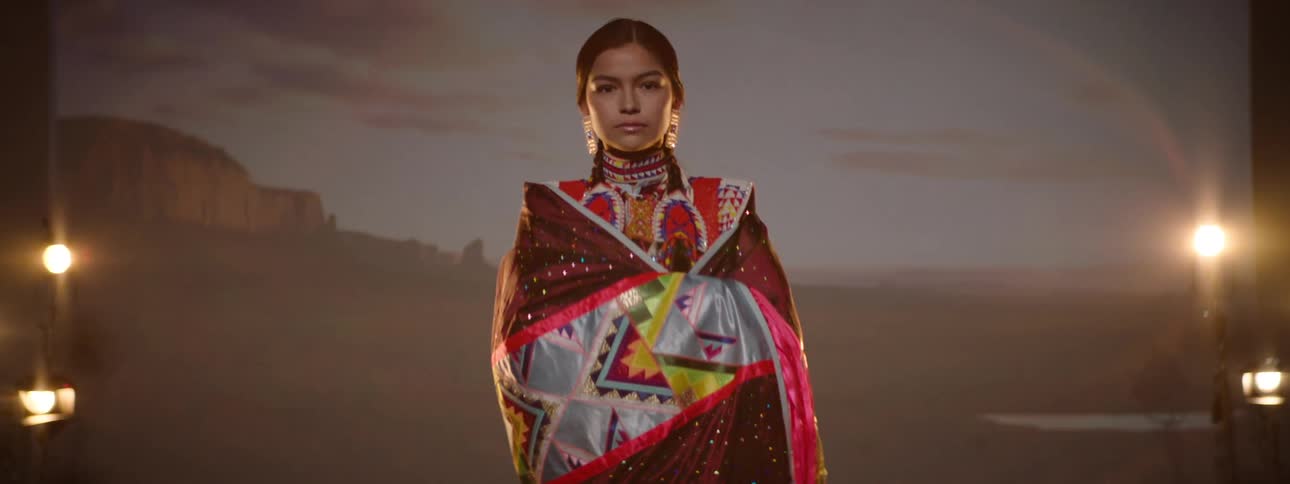 A woman in indigenous regalia spreads a fringed shawl under stage lights
