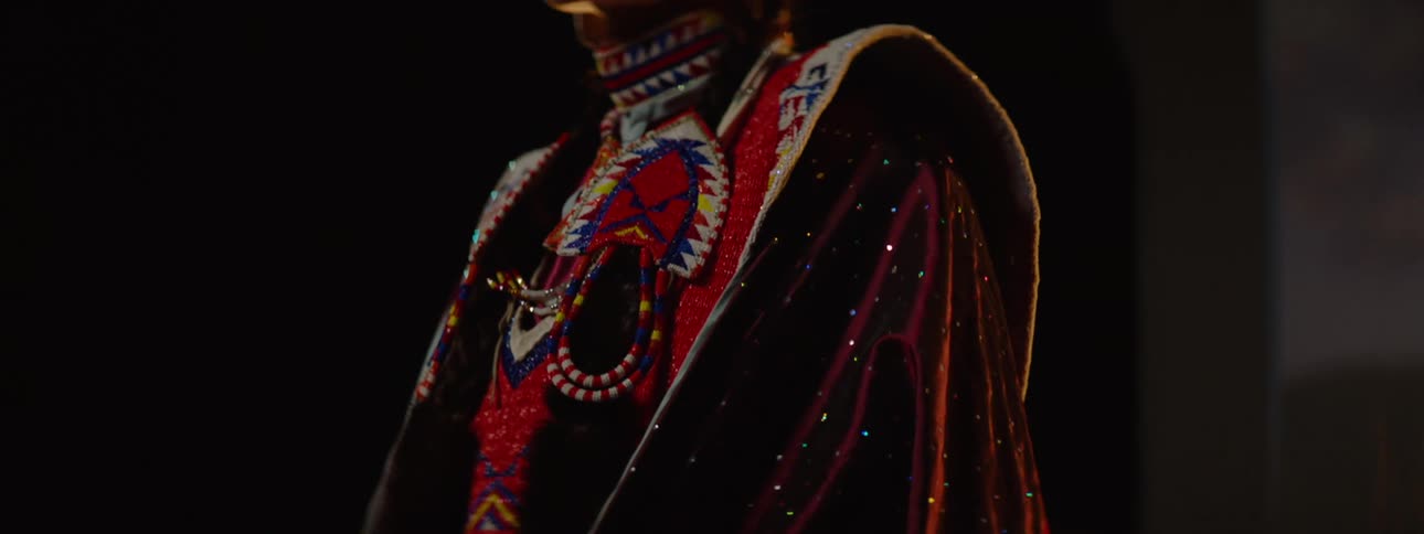 A person wearing vibrant intricately beaded and patterned traditional indigenous regalia is seen gleaming under soft light in a dark setting