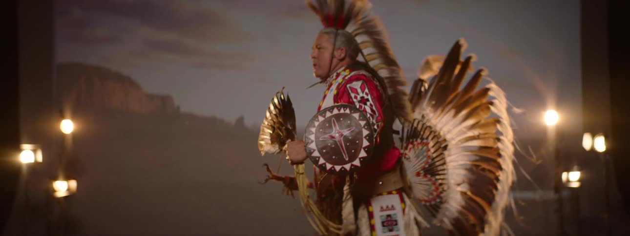 A man in elaborate traditional indigenous native american regalia featuring a feather headdress and bustle holds a feathered staff and a decorated shield while performing against a warm blurred background