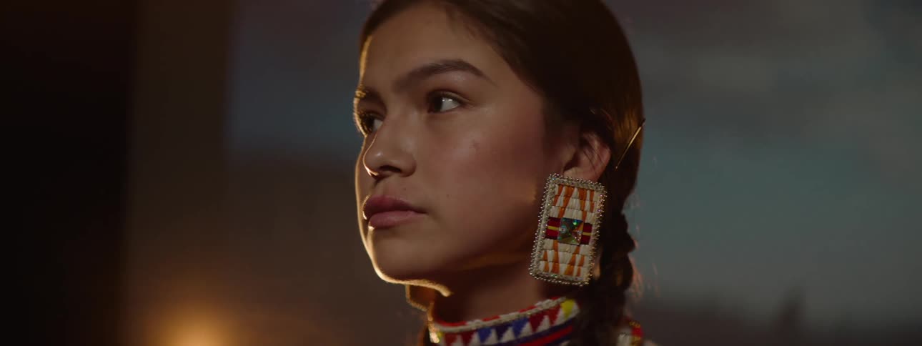 A native american teenage girl wearing traditional attire adorned with jewelry and makeup stands confidently on a stage