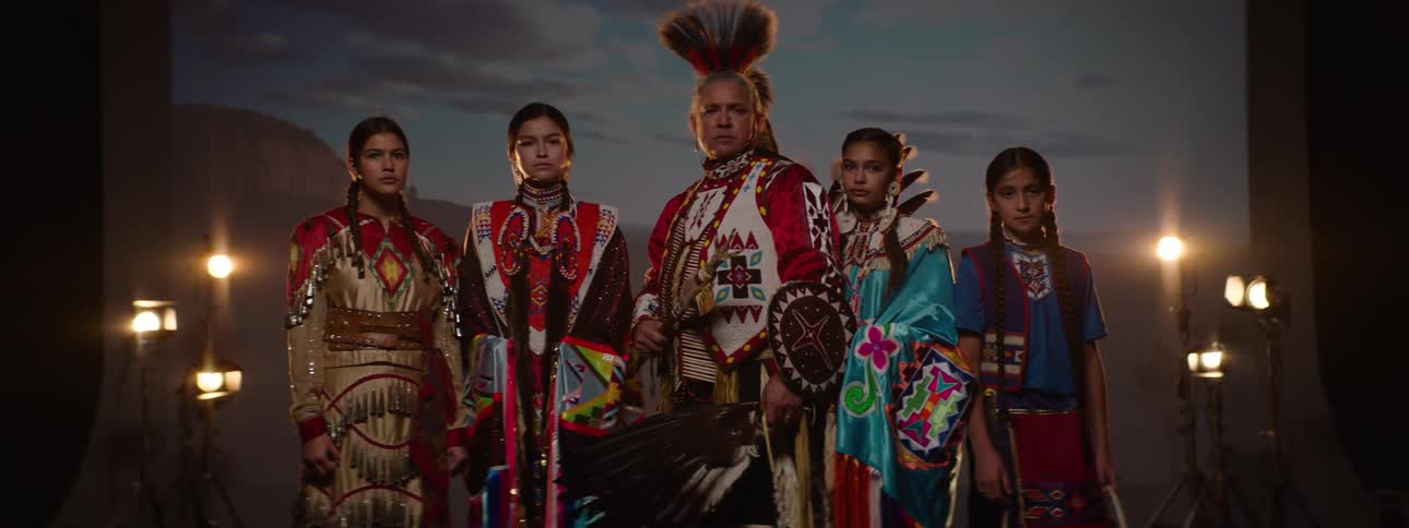 Group of five people in traditional indigenous costumes posing side by side under stage lighting