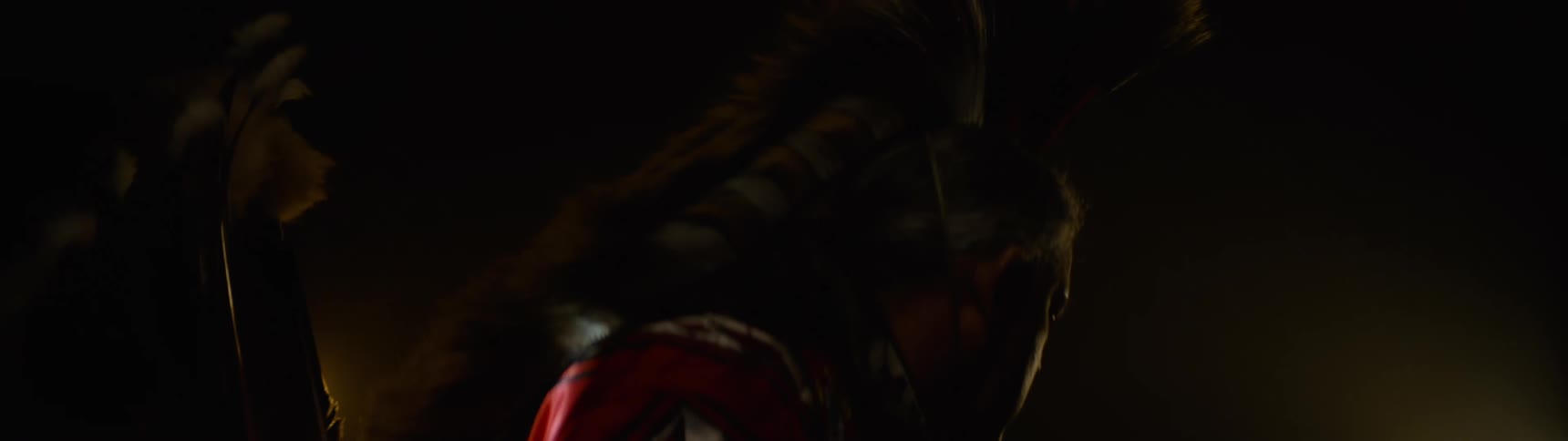 A person wearing traditional indigenous native american regalia with red and white patterns and a feathered headdress is seen in profile against a dark background illuminated by a warm low light source from the left