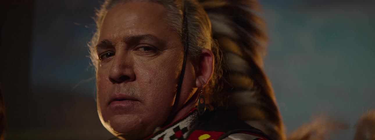 Older male dancer stands in native american ceremonial attire near a photography backdrop