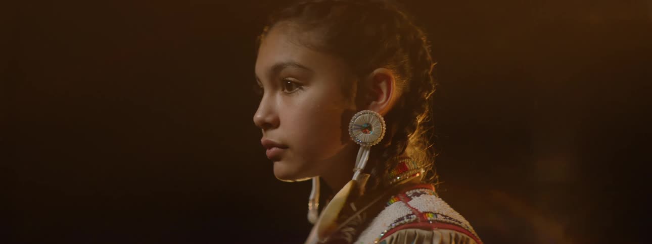 A teenage native american girl wearing traditional attire stands on a stage