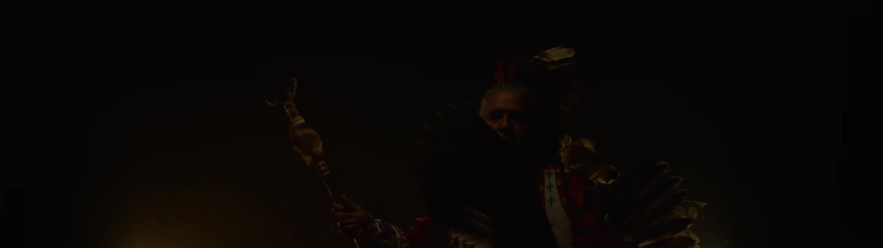 A native american person wearing a feathered headdress and traditional regalia dances in a dark hazy environment while holding a tomahawk and looking directly at the camera with a bright light source behind them