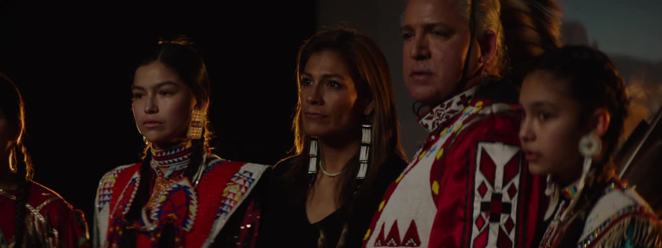 A group of native american individuals are standing sidebyside in traditional regalia against a dark background