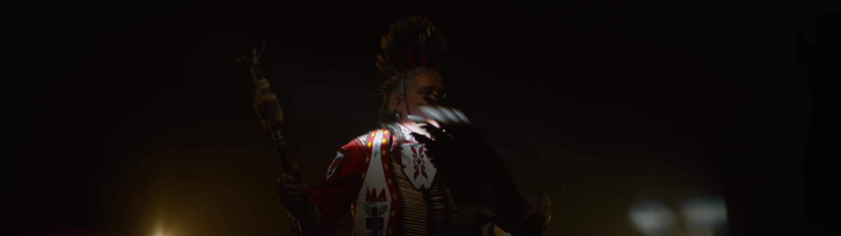 An individual in elaborate native american ceremonial attire featuring large feathers and intricate details is dimly lit in a dark hazy environment with bright lights visible on either side