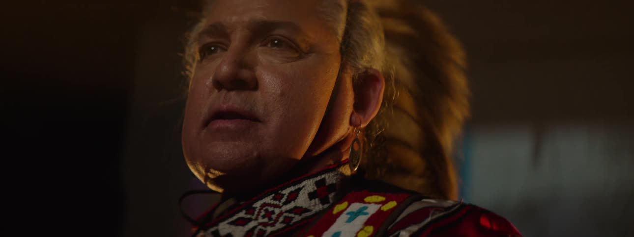 Older man wearing native american ceremonial attire stands in a room lit by electric lights