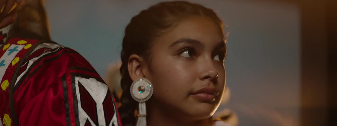 A teenage native american girl stands next to a man on a stage both wearing traditional native attire