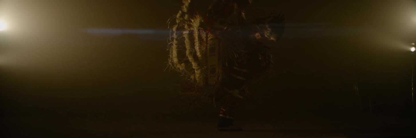 Male dancer wearing traditional native american ceremonial attire dances in a dim room lit by small electric lights