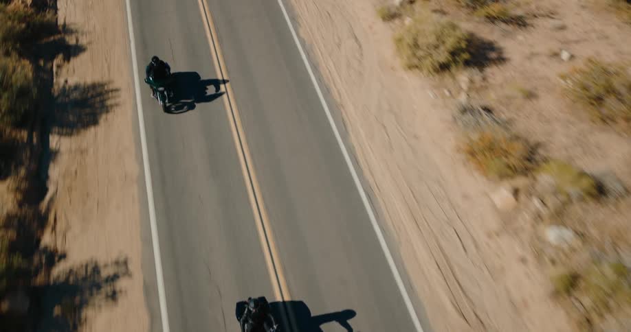 An aerial shot captures two motorcycles casting long shadows as they travel on a twolane road that cuts through a sunlit arid landscape