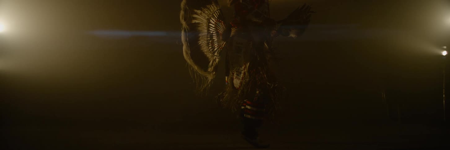 A figure in elaborate feathered indigenous regalia dances slowly in a dimly lit hazy space their attire highlighted by a strong backlight that creates a prominent horizontal lens flare