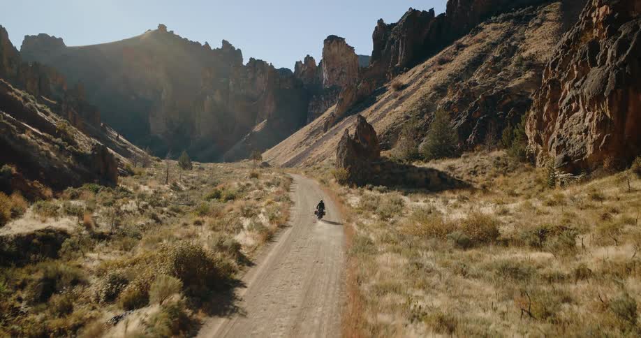 A person wearing all black rides a motorcycle down a path between two mountains