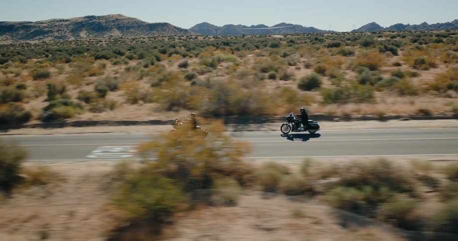 Low aerial view of two motorcycles driving down a desert highway near hills in the background on a bright day