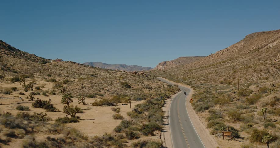 Two motorcyclists ride down a winding desert road surrounded by dry hills
