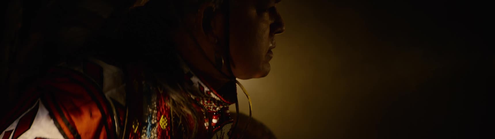 Spotlight moves across an older male dancer performing in native american ceremonial attire