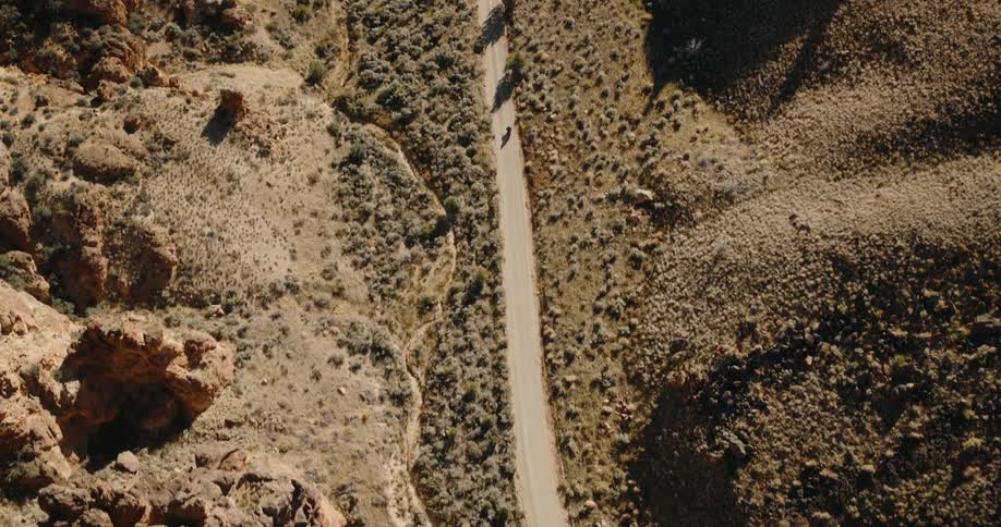 A lone person rides a bike down a narrow dirt road cutting through a rocky canyon landscape
