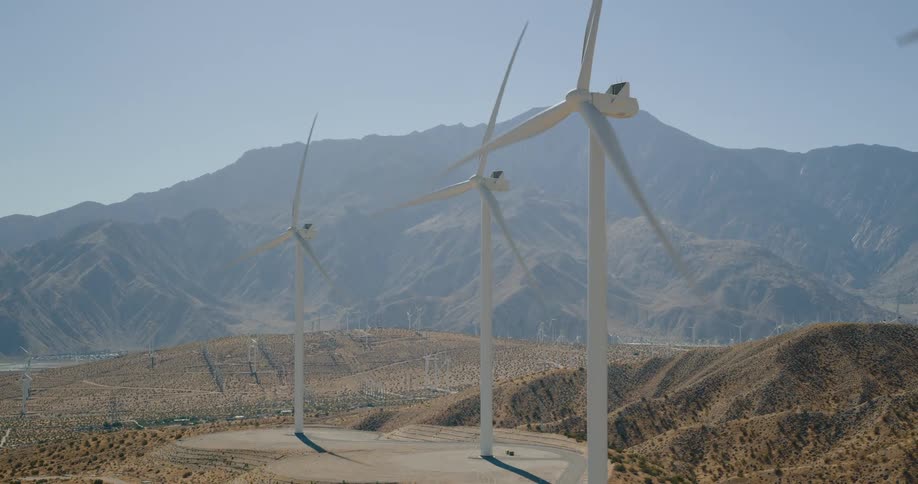 Large wind turbines tower above a dry desert valley with rugged mountains in the distance