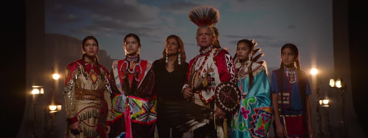 A group of american indians including a man a woman and several teenage girls stand together on a stage dressed in traditional native attire