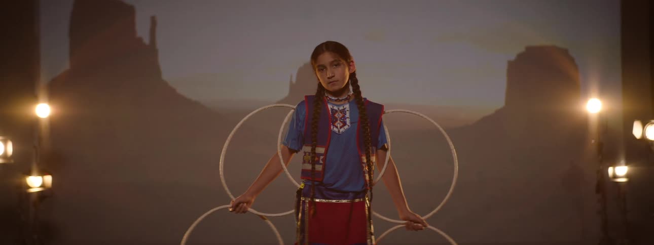 Young girl in traditional attire performs hoop dance on dim stage with canyon backdrop