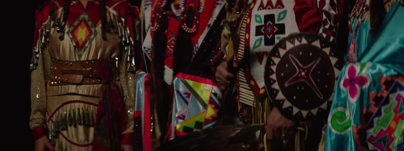 A row of native american individuals in traditional regalia featuring vibrant patterns and feathered elements stands in a dark setting with the person in the center appearing to be a man with a stern expression