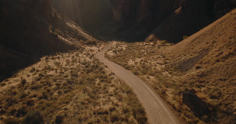 An aerial view pulls back along a winding dirt road that carves through a vast sunlit canyon with dry rugged hillsides