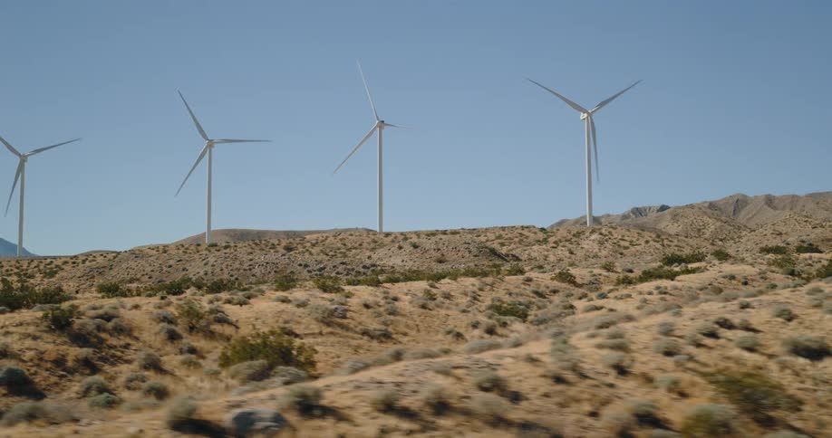 White wind turbines stand on a dusty rocky hill covered in sparse green vegetation under a clear blue sky