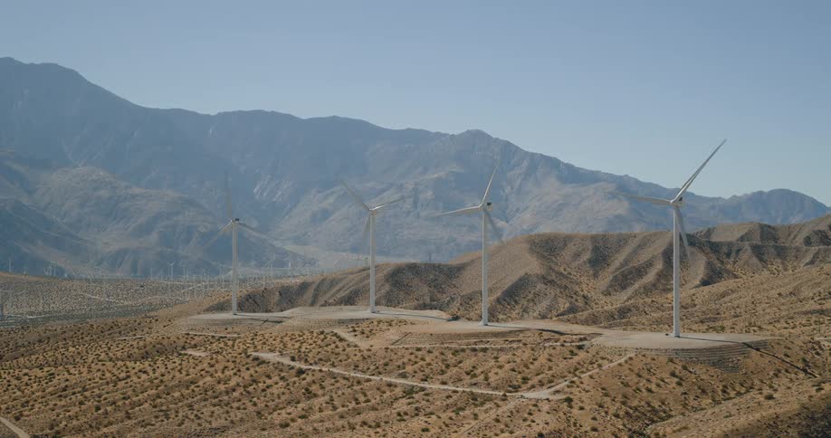 An aerial view pulls back from a row of wind turbines standing across open fields revealing a vast rural landscape under soft daylight