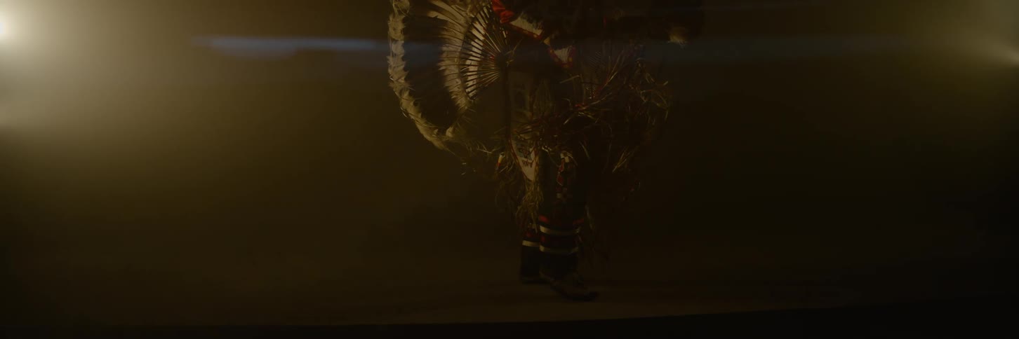 A man dances in elaborate native american ceremonial attire adorned with large feathers and intricate details in a dark hazy environment with bright lights on either side