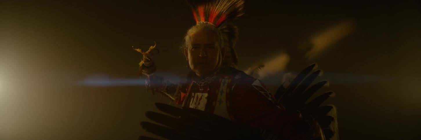 A native american man holding a tomahawk and looking directly at the camera is wearing a feathered headdress and traditional regalia in a dark hazy environment with a bright light source